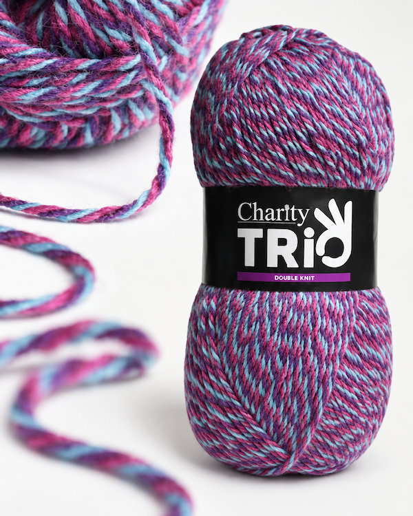 A skein of Charity Trio double knit yarn in a variegated blend of purple, pink, and blue, with a matching ball of yarn and loose strands blurred in the background on a white surface.